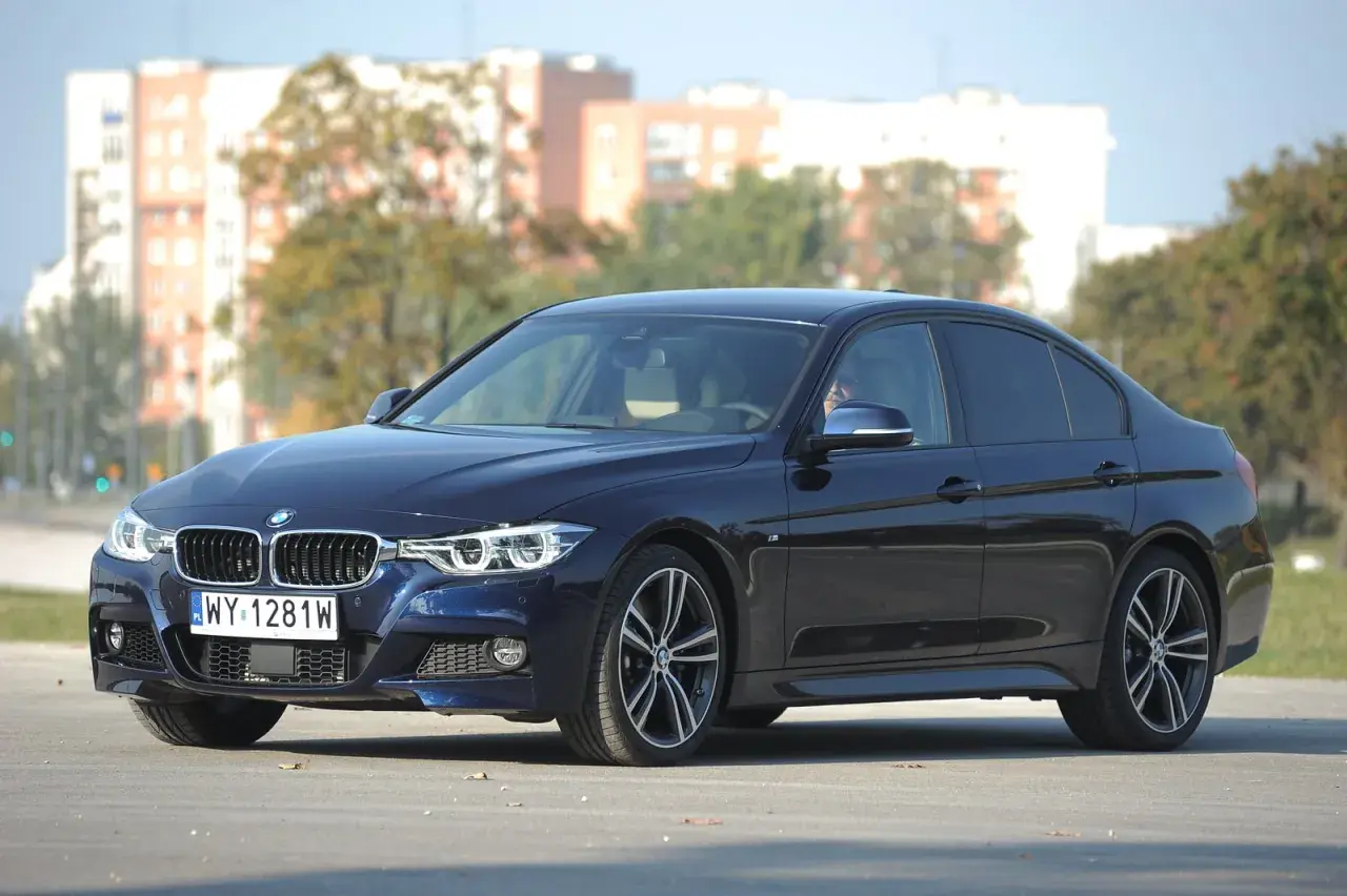 Ciemnoniebieskie BMW 318d z polskimi tablicami rejestracyjnymi stoi na tle budynków i drzew.