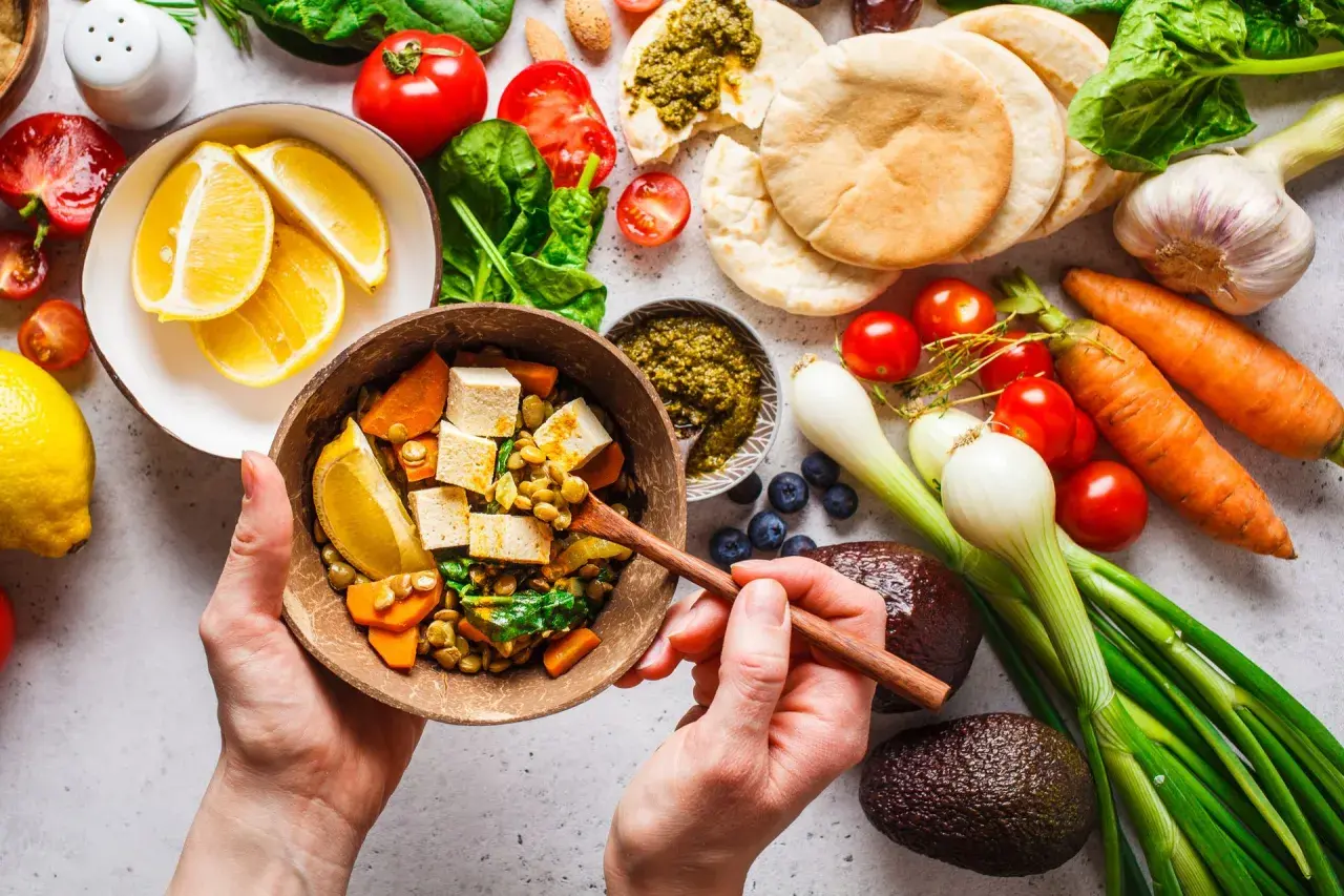 Zdrowa dieta na stawy: miska pełna soczewicy, tofu, warzyw i cytryny. Obok składniki na pyszne danie: pomidory, marchew, awokado, czosnek, cebula, jagody, pesto i chlebki pita.