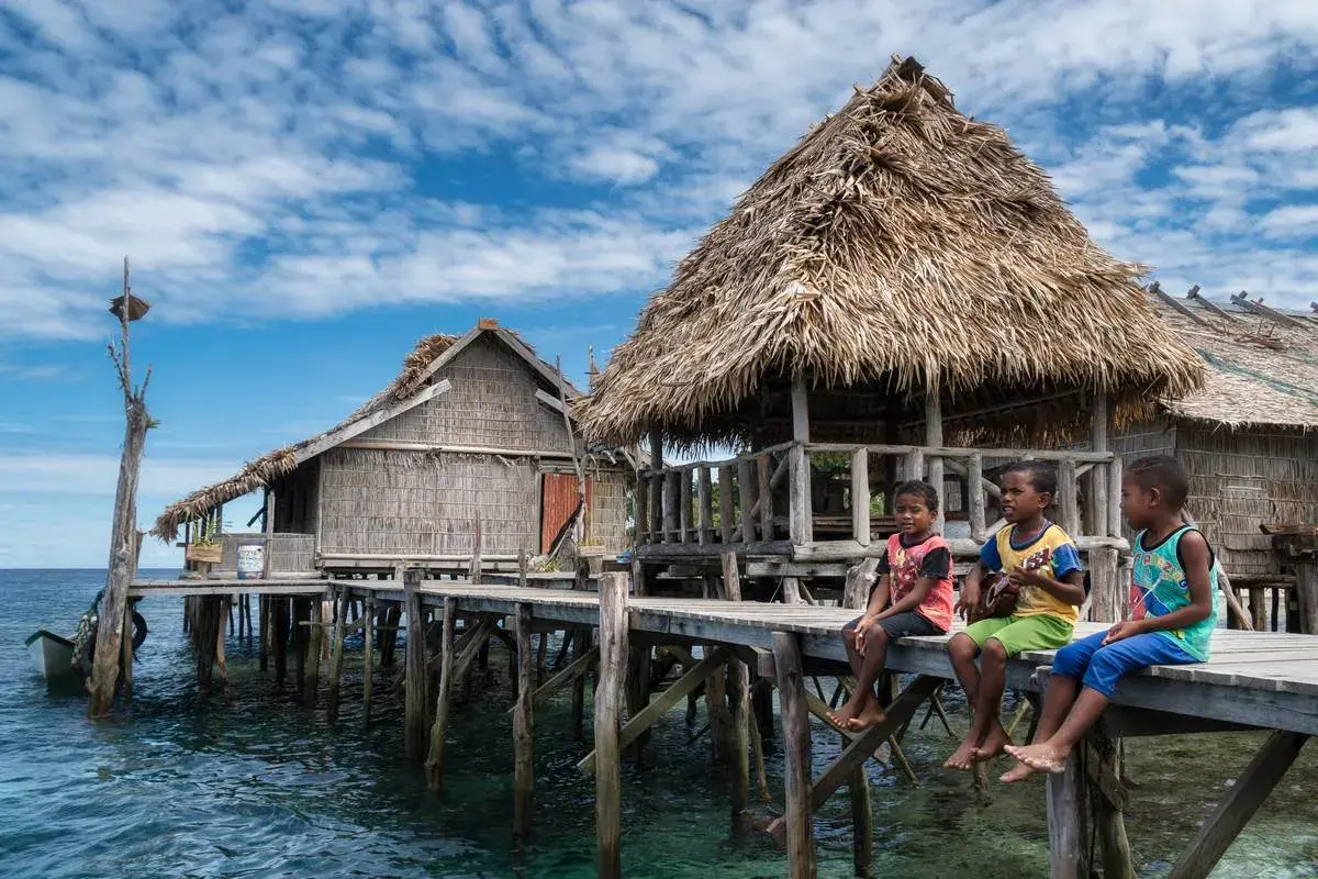 Trzech chłopców siedzi na drewnianym pomoście przy tradycyjnych chatach na wodzie w Raja Ampat.