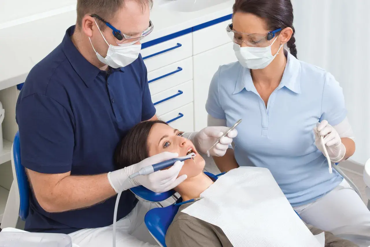 Dentysta i asystentka w okularach i maseczkach pracują przy pacjentce. Asystentka trzyma narzędzie, a dentysta używa **vector paro** do zabiegu.