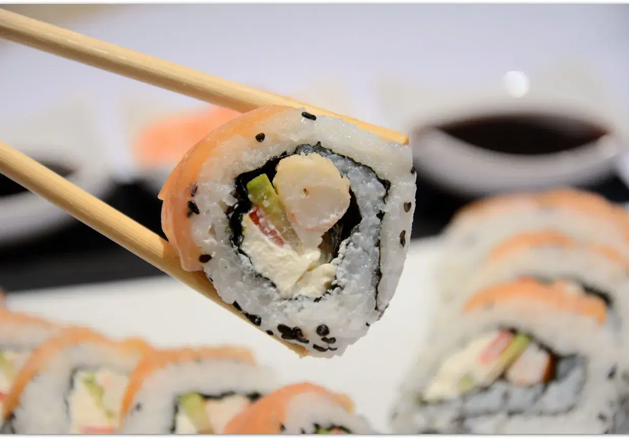 Chopsticks trzymają sushi z krewetkami, awokado i kremowym serkiem. W tle widać więcej roladek sushi.
