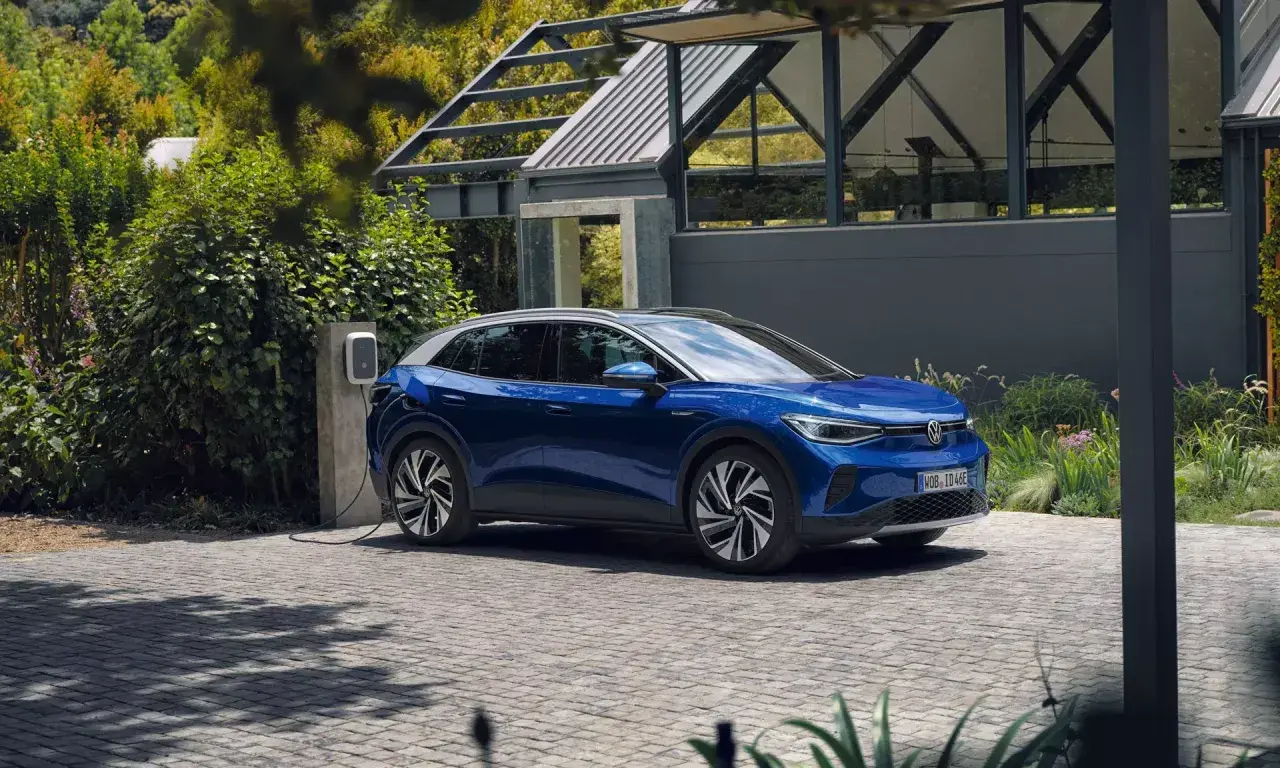 Niebieski Volkswagen ID.4 ładuje się przy domu, otoczony zielenią.