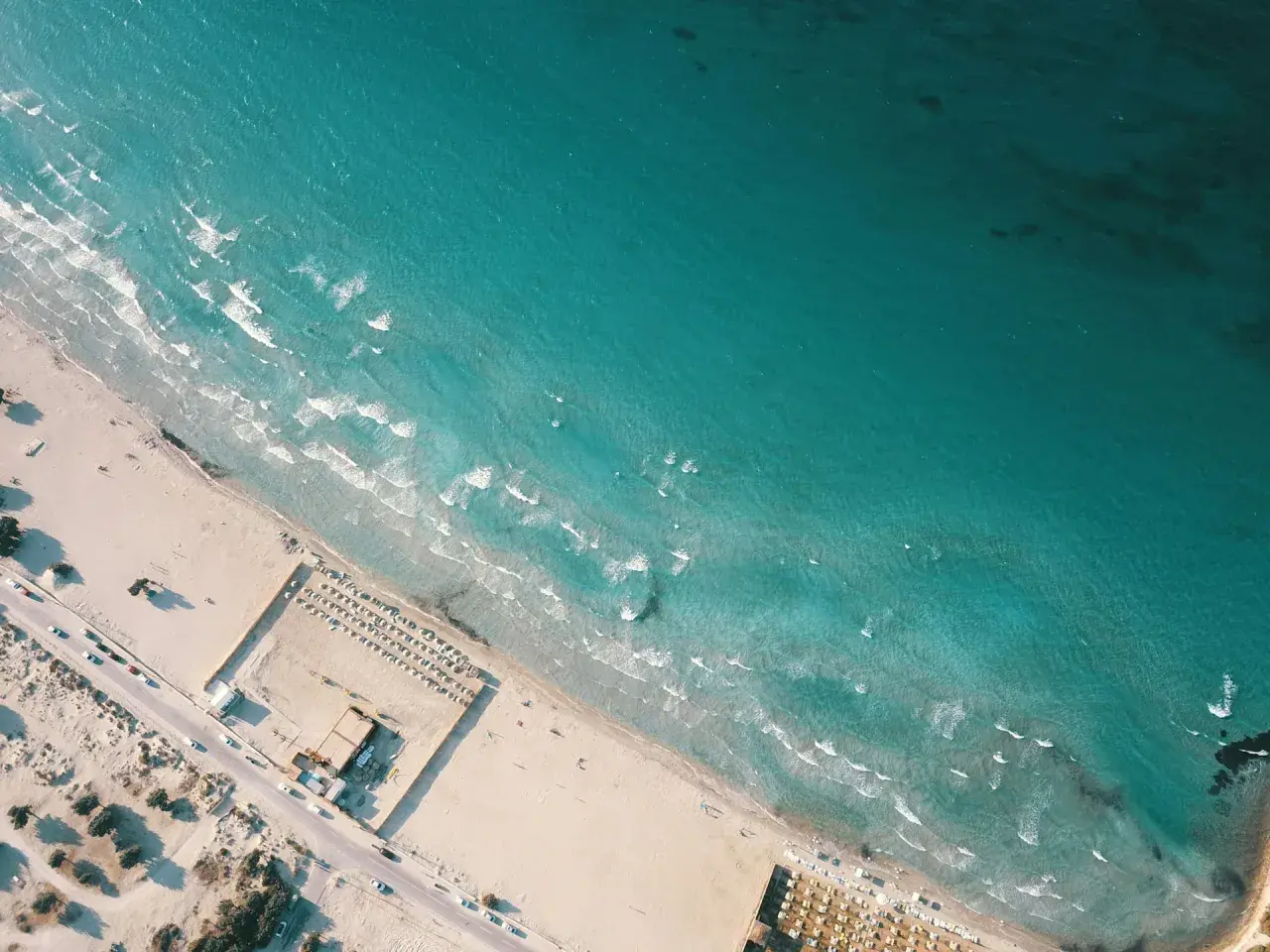 Izmir nad Morzem Egejskim. Piękna, piaszczysta plaża z turkusową wodą i falami. W oddali widać leżaki i parasole.