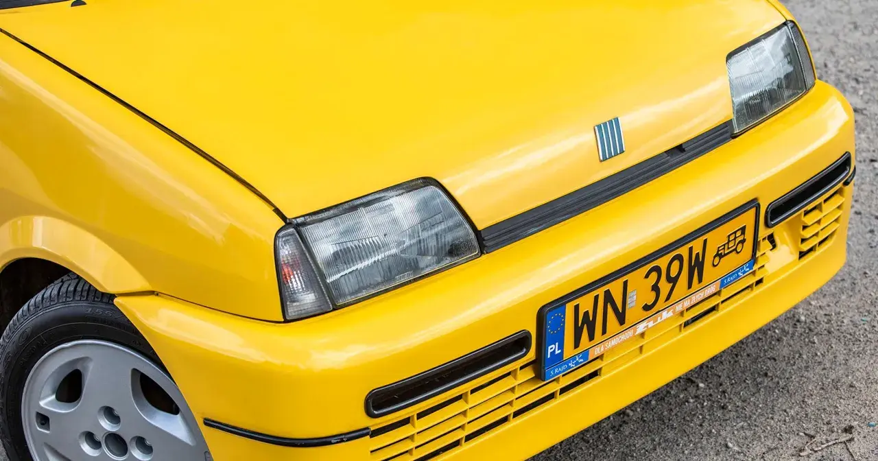 Żółte tablice rejestracyjne na błyszczącym, żółtym samochodzie Fiat. Widoczny fragment maski, reflektor i zderzak.