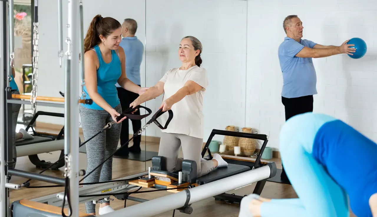 Frauen und Männer trainieren im **Reha-Sport** auf Pilatesgeräten. Eine Trainerin hilft einer älteren Frau, während ein Mann mit einem Ball übt.