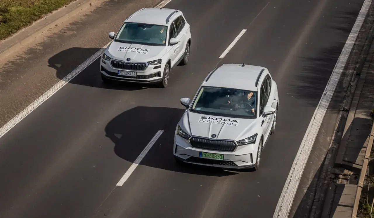 Dwa białe samochody Skoda Auto Szkoła jadą po drodze, gdzie obowiązuje ruch lewostronny.