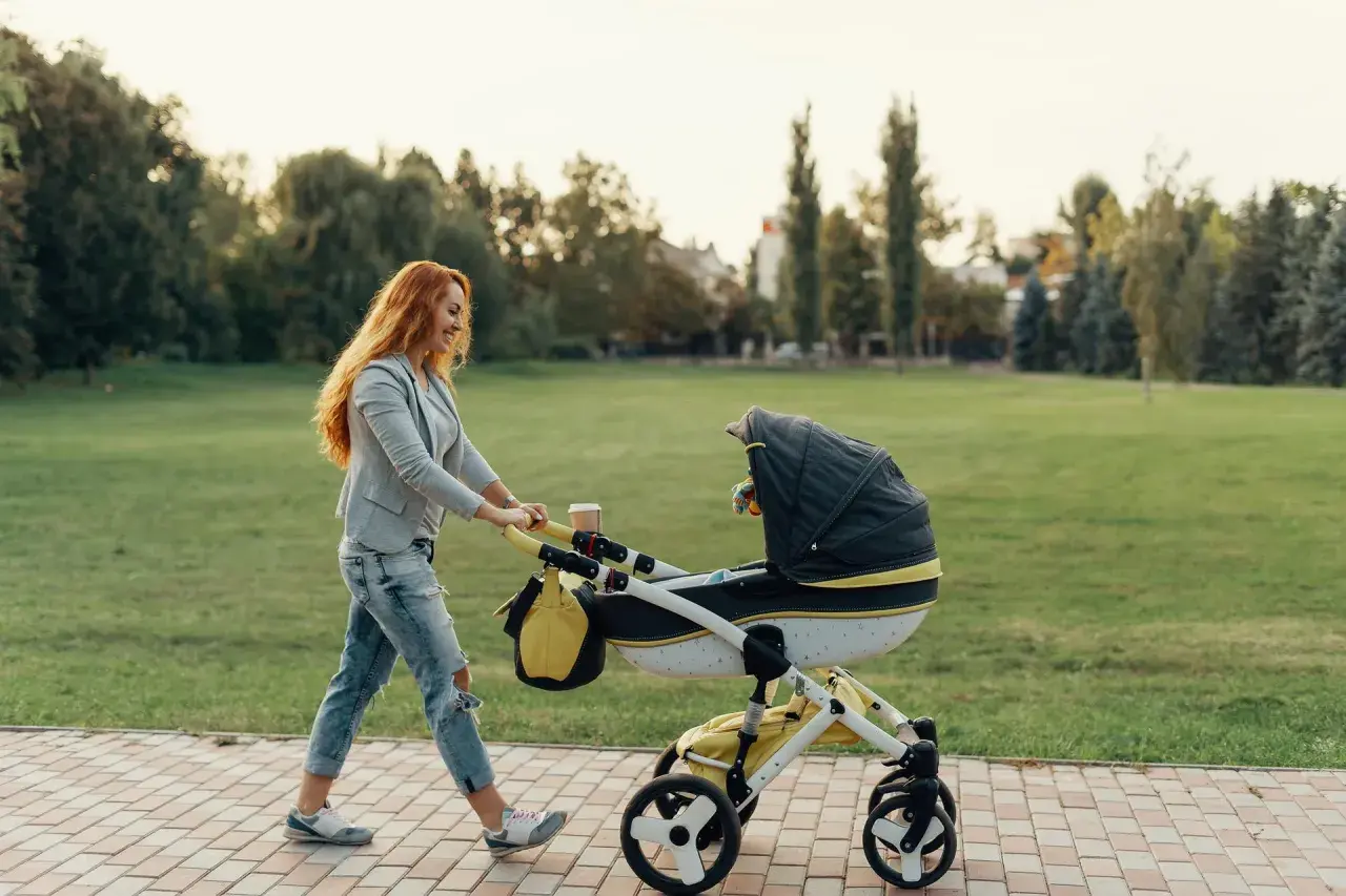 Ruda kobieta z uśmiechem pcha wózek podczas pierwszego spaceru z noworodkiem w parku.