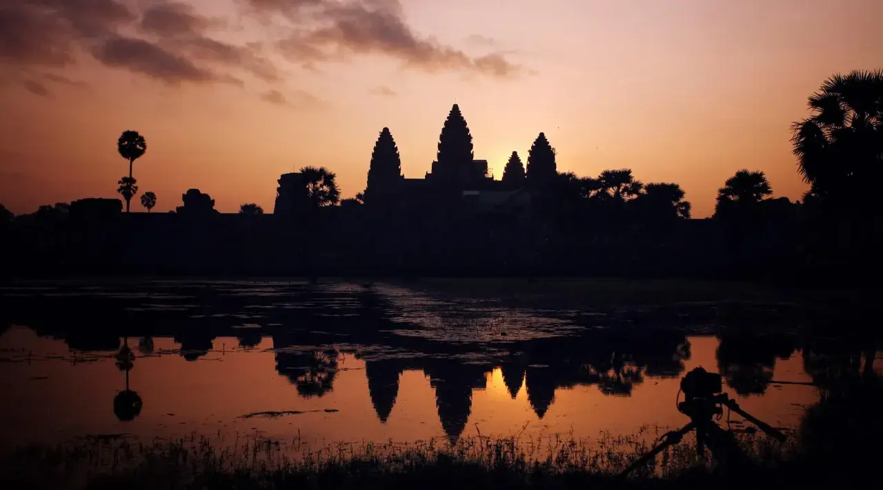 Zachód słońca nad Angkor Wat, idealne tło dla niezapomnianych kambodżańskich wakacji. Widok odbija się w wodzie, a na pierwszym planie statyw z aparatem.