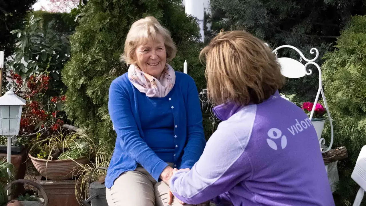 Eine freundliche ältere Dame im Gespräch mit einer Pflegekraft. Deren Kleidung trägt das Logo "vidon". Ein Gutachter für den Pflegegrad könnte hier im Gespräch sein.