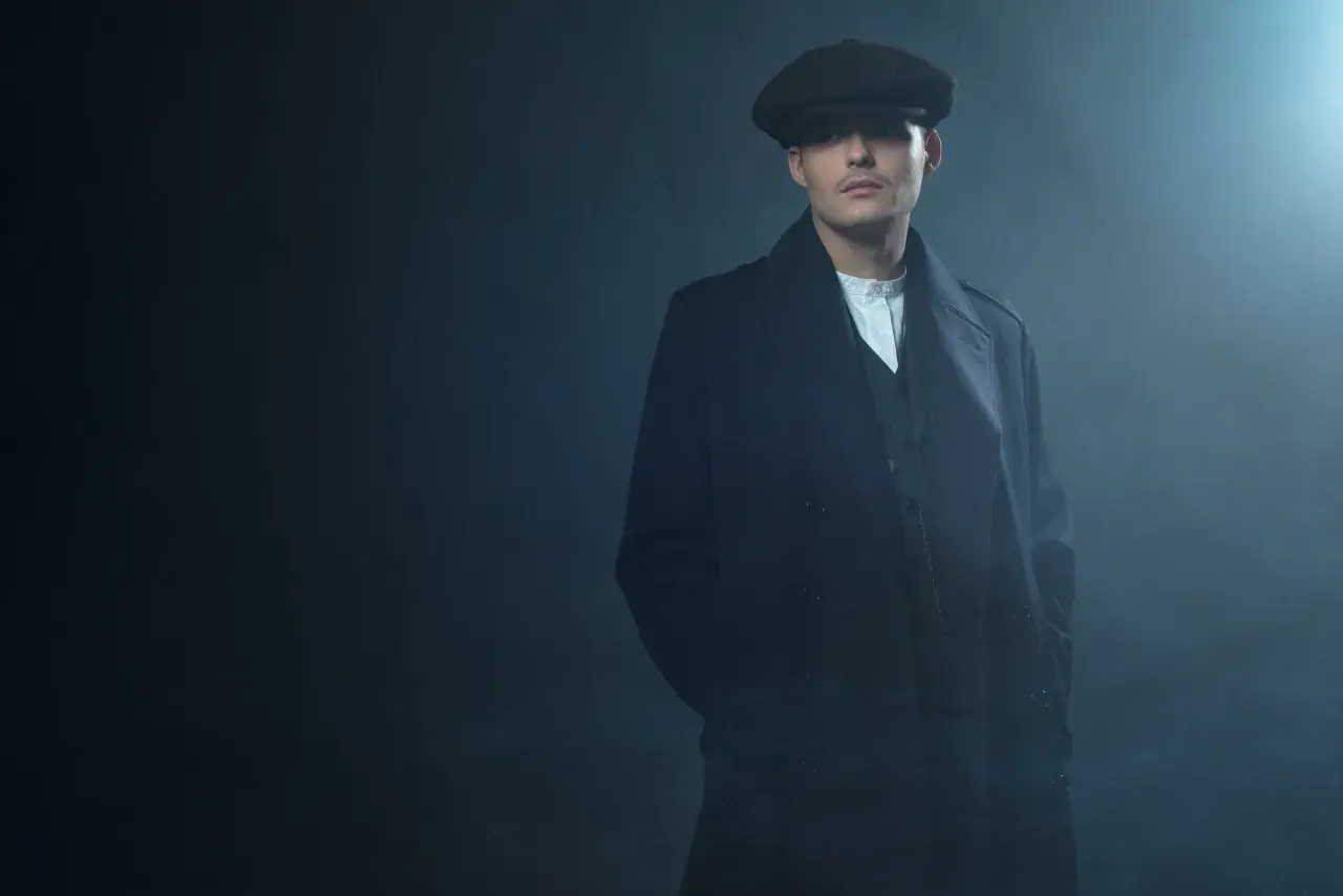 Mężczyzna w kaszkiecie i płaszczu, z charakterystycznym wąsem, emanuje chłodnym spojrzeniem w stylu Peaky Blinders.