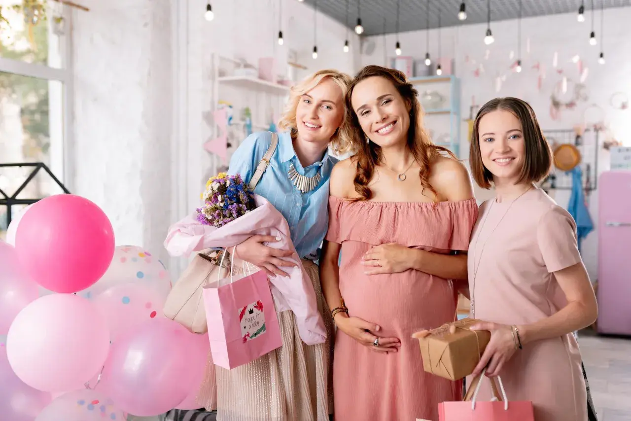 Trzy uśmiechnięte kobiety świętują baby shower. Jedna z nich jest w ciąży, a pozostałe dwie trzymają prezenty i balony.