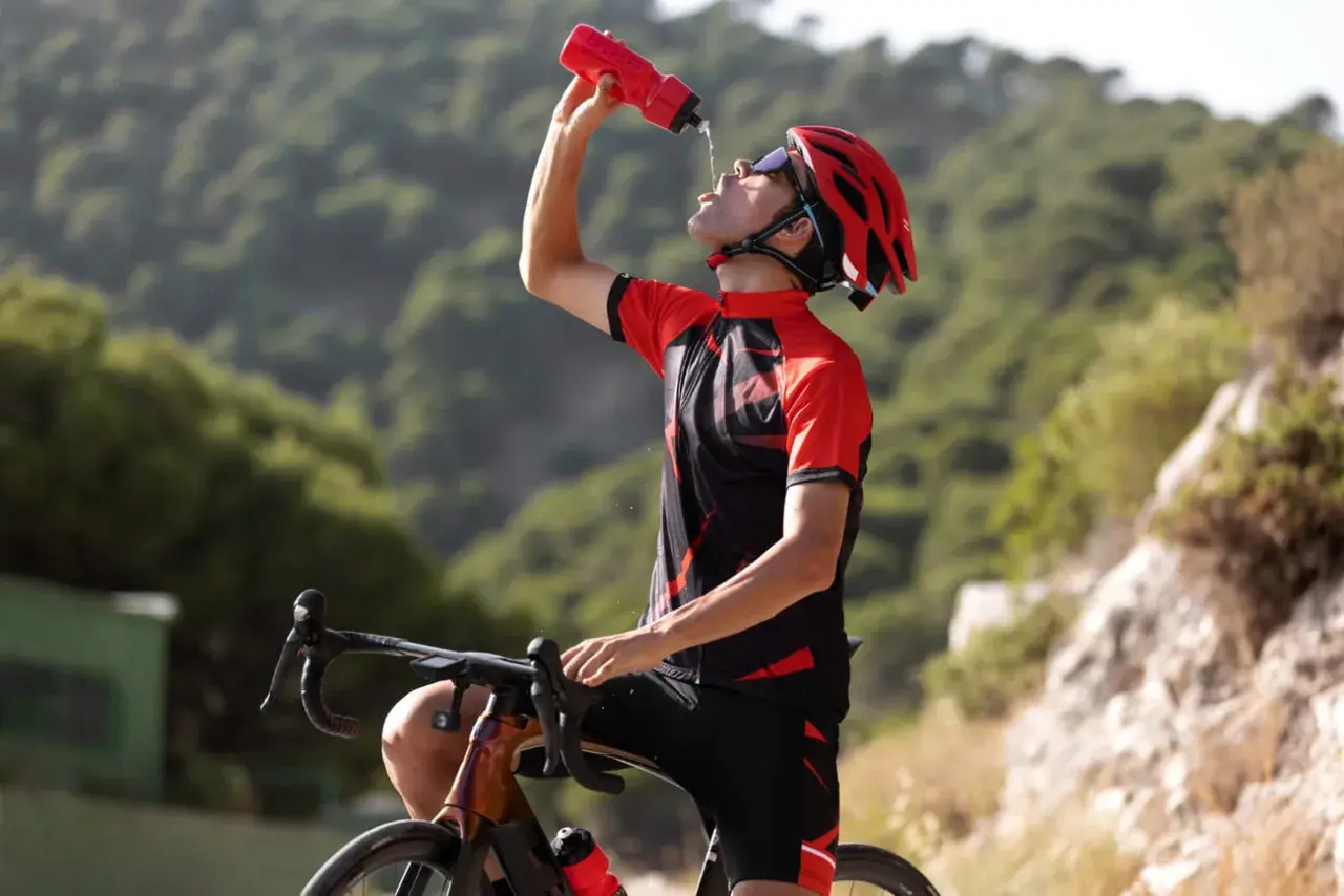 Kolarz w kasku i stroju sportowym pije wodę z bidonu, regenerując siły po wysiłku. Woda z izoleucyną pomoże mu szybciej odzyskać energię.
