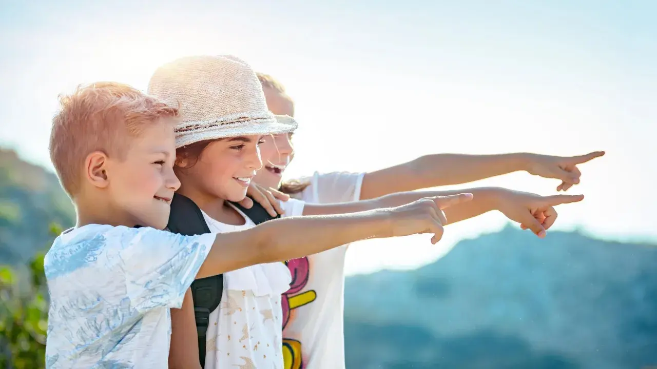 Trois enfants souriants, portant des sacs à dos, pointent du doigt vers la droite, sous un ciel ensoleillé.