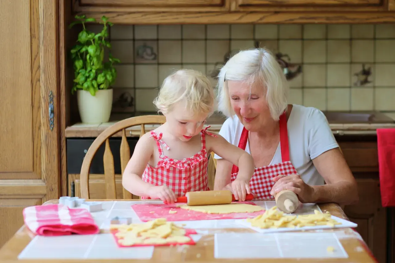 Babcia z wnuczką pieką ciasteczka. Dziewczynka wałkuje ciasto, a babcia z uśmiechem patrzy na jej pracę.