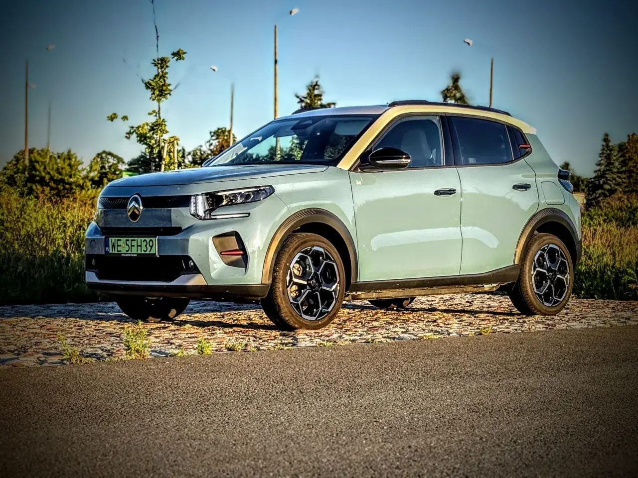 Jasnoszary Citroën ë-C3 z żółtym dachem stoi na tle drzew i nieba.