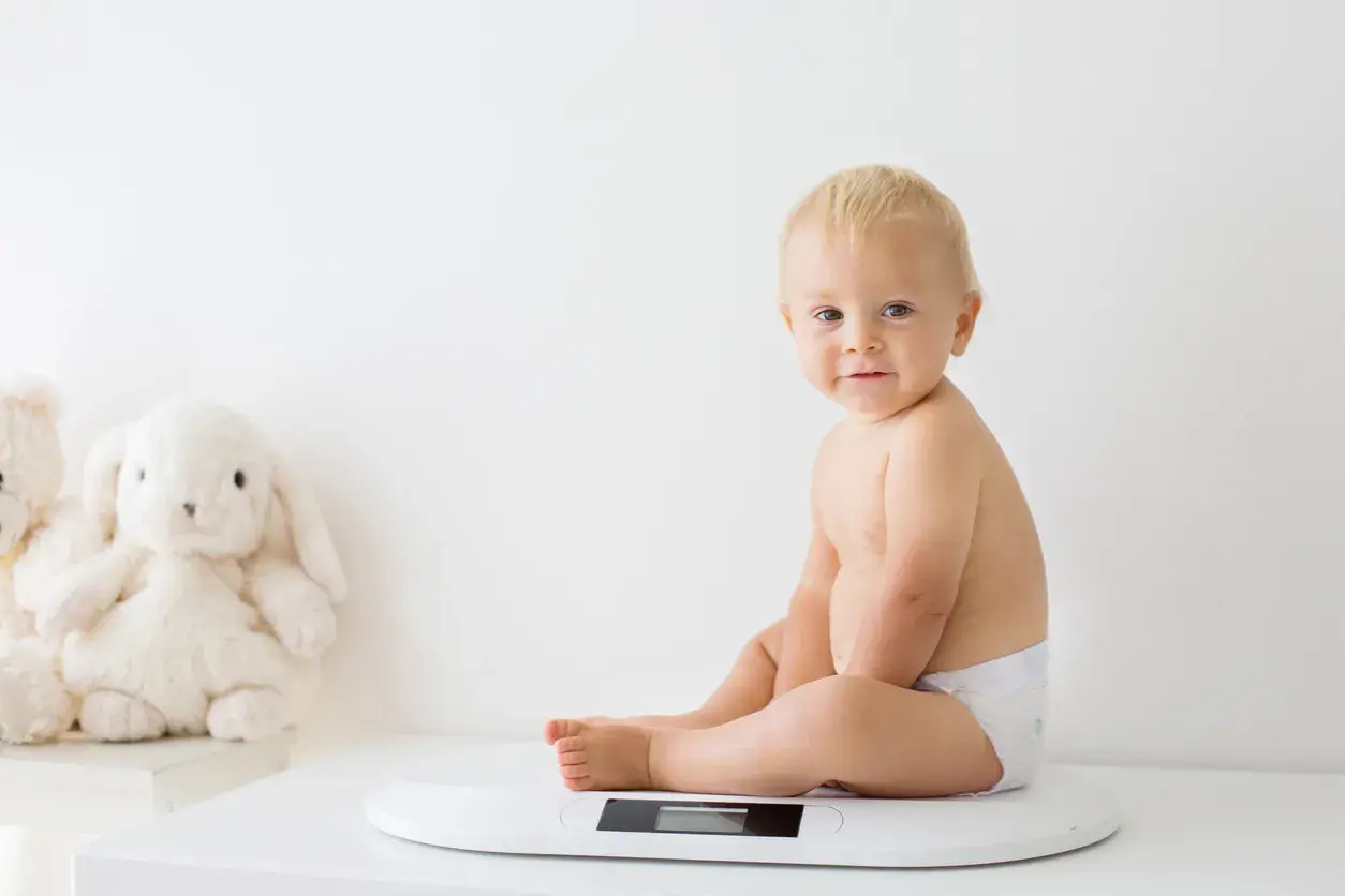 Uśmiechnięty chłopczyk w pieluszce siedzi na wadze. Chyba już czas na większy rozmiar, kiedy pampers jest za mały.