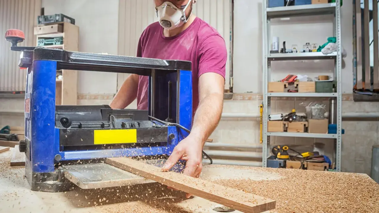 Stolarz w masce ochronnej pracuje przy strugarce, wygładzając drewno. Wióry sypią się wokół.