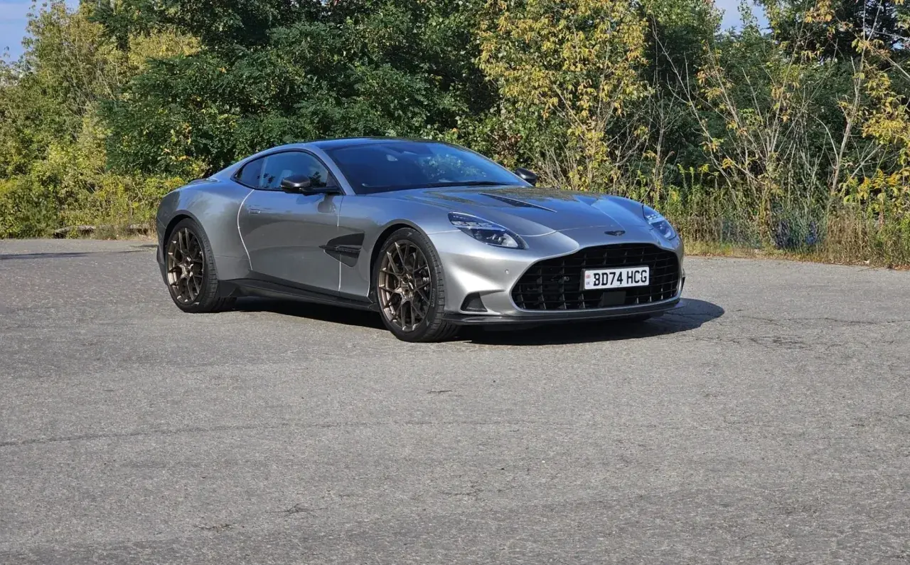 Srebrny Aston Martin Vanquish 2025 z brązowymi felgami na tle zieleni.