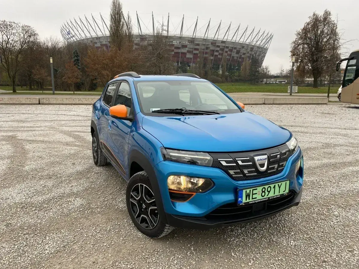 Niebieski Dacia Spring z pomarańczowymi akcentami na tle Stadionu Narodowego. Sprawdź atrakcyjną dacia spring cena!