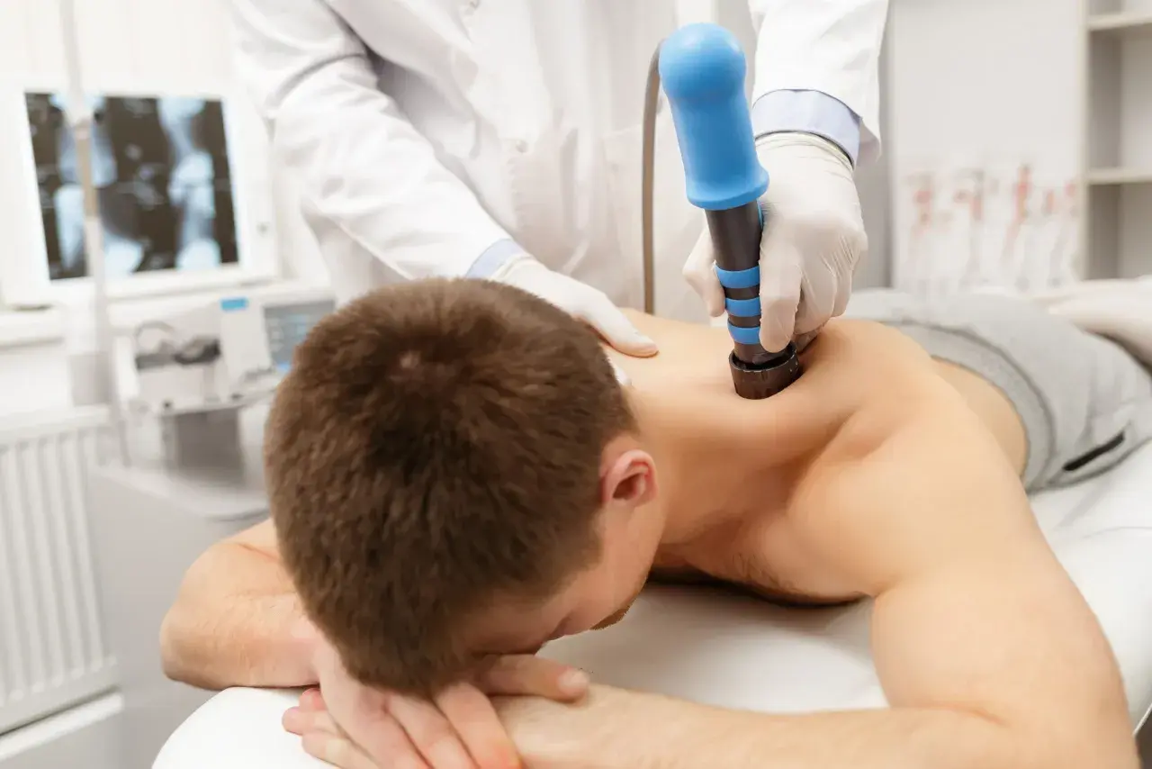 Arzt behandelt HWS-Syndrom mit Stoßwellentherapie. Ein Mann liegt auf dem Bauch, während ein Arzt mit einem blauen Gerät auf seinen Rücken drückt.