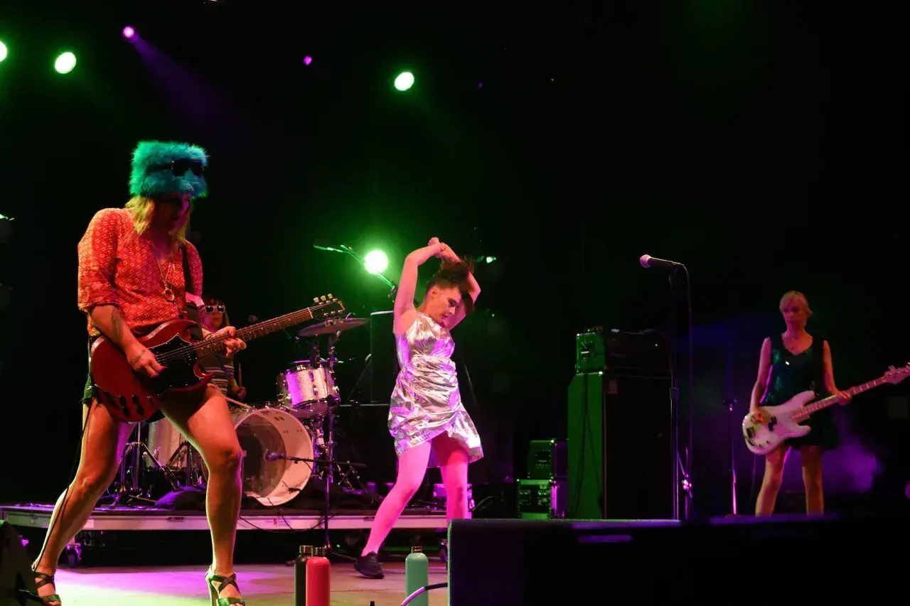 Sur scène, le groupe Bikini Kill enflamme le public. Une guitariste porte un chapeau vert fluo, une autre joue de la basse, et une troisième danse avec énergie.