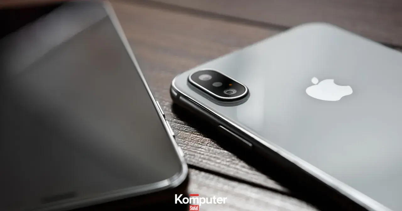Dwa telefony leżą obok siebie na drewnianym blacie. Jeden z nich, srebrny iPhone, ma widoczny moduł aparatu. Drugi, z ciemnym ekranem, jest ustawiony bokiem. Idealne do rozmowy przez **Facetime**.