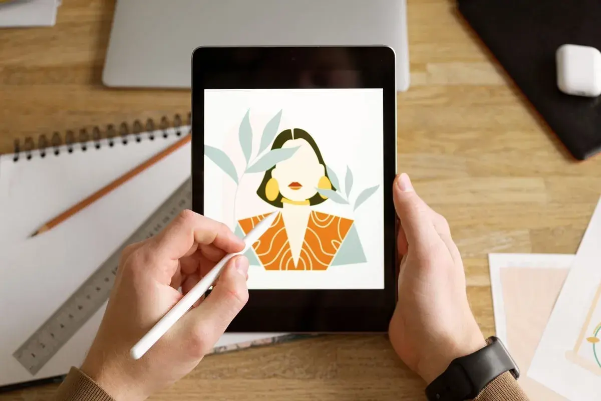 Ręce trzymają tablet z rysunkiem kobiety, tworzonym w aplikacji procreate. Obok leżą szkicownik i ołówek.