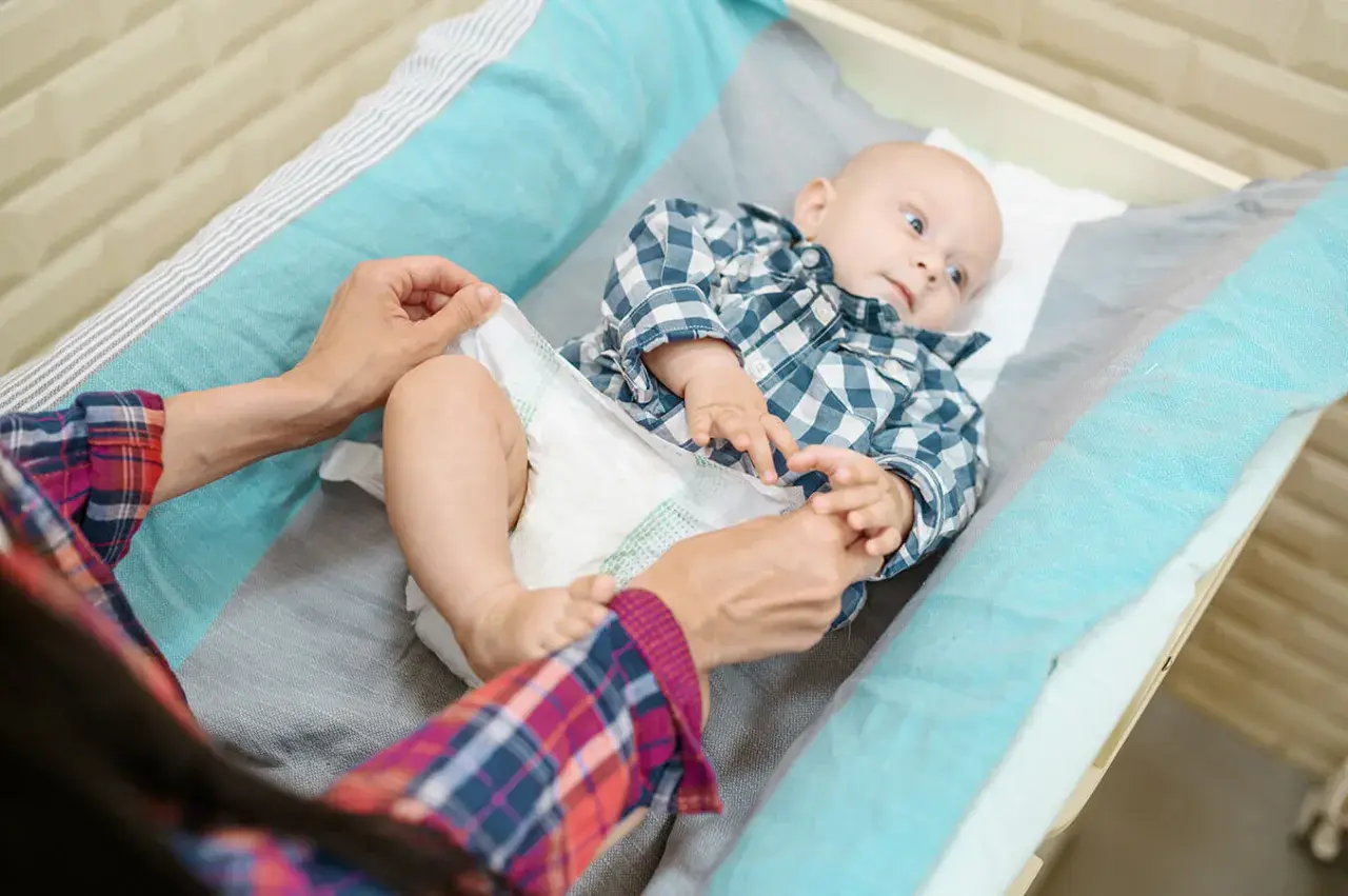 Mama sprawdza, kiedy zmienić rozmiar pieluchy maluszkowi. Dziecko leży na przewijaku w kratkę.