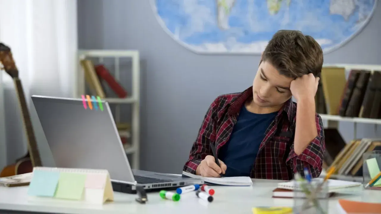 Ein Junge sitzt frustriert am Schreibtisch und macht seine hausaufgaben. Ein Laptop und bunte Stifte liegen bereit.