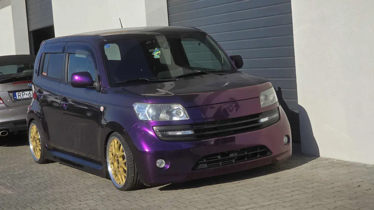 Fioletowy Daihatsu Materia z błyszczącymi złotymi felgami, zaparkowany przed garażem.