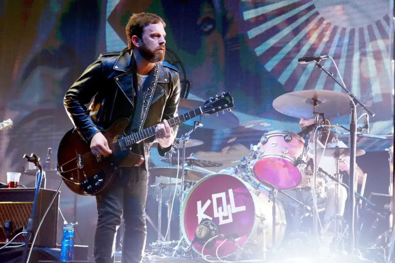 Gitarzysta Kings of Leon Polska w skórzanej kurtce gra na scenie. W tle perkusja z logo KQL.