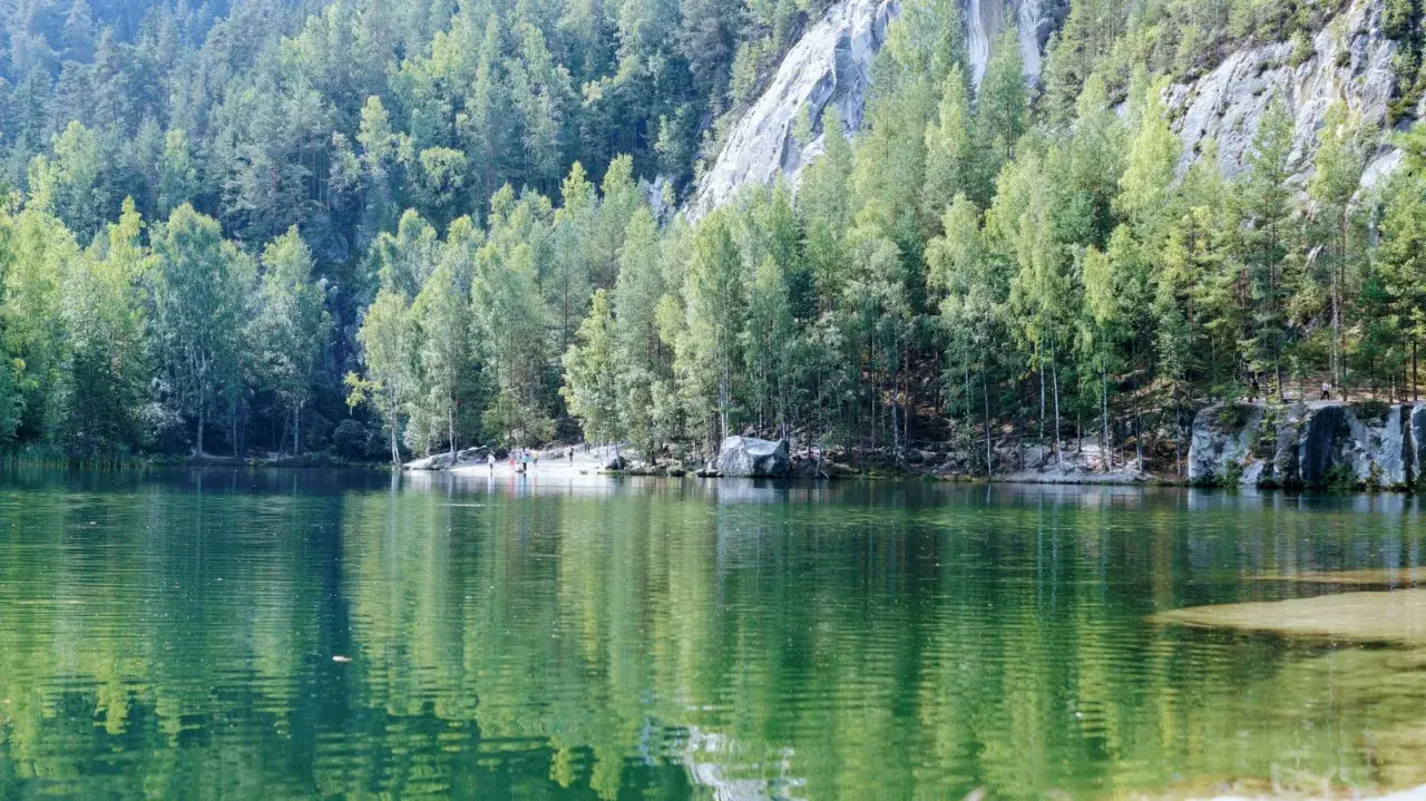 Zielona tafla jeziora odbija las i skalne miasto w Polsce. Ludzie spacerują po piaszczystym brzegu.