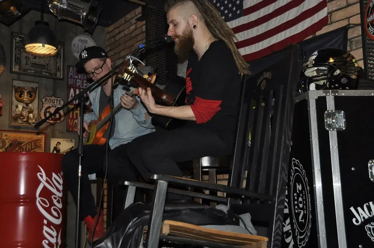 Dwóch muzyków gra na gitarach w klimatycznym wnętrzu. Jeden z nich, muzyk grający do kotleta, ma dredy i brodę.