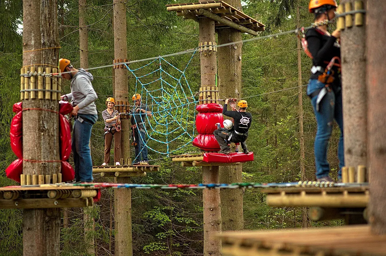 Park linowy Base Camp Istebna: ludzie na platformach wśród drzew, pokonują przeszkody, w tym pajęczynę.