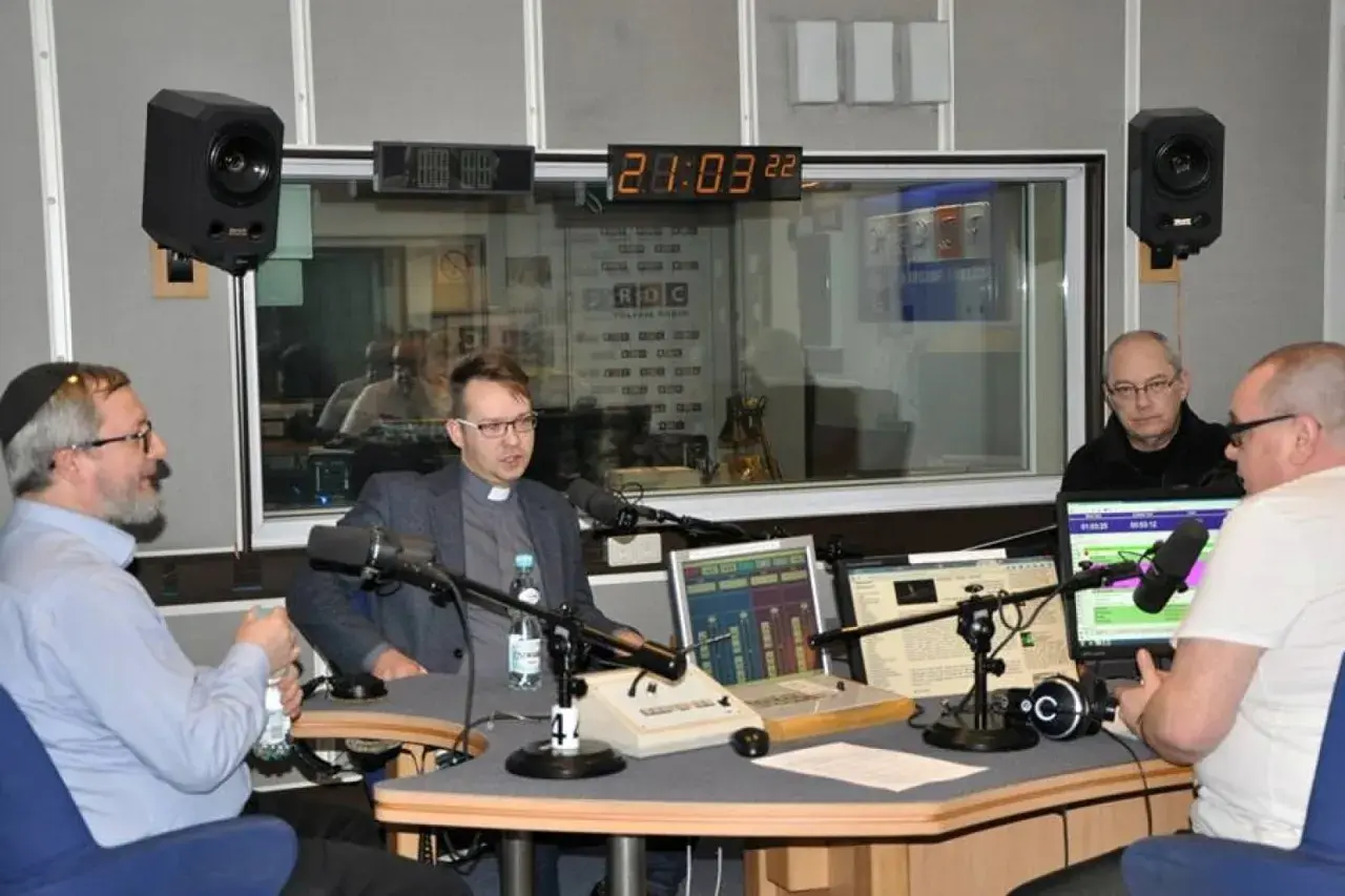 W studiu radiowym czterech mężczyzn, w tym ksiądz i osoba w jarmułce, rozmawia przy mikrofonach. W tle widać zegar i logo RDC.