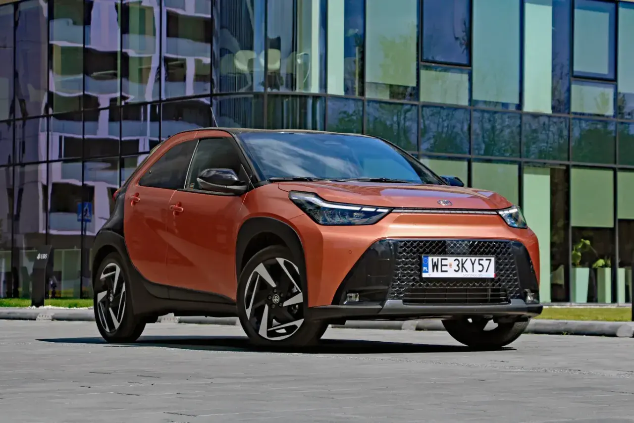 Pomarańczowy Toyota Aygo X Hybrid z czarnymi detalami stoi przed nowoczesnym budynkiem.
