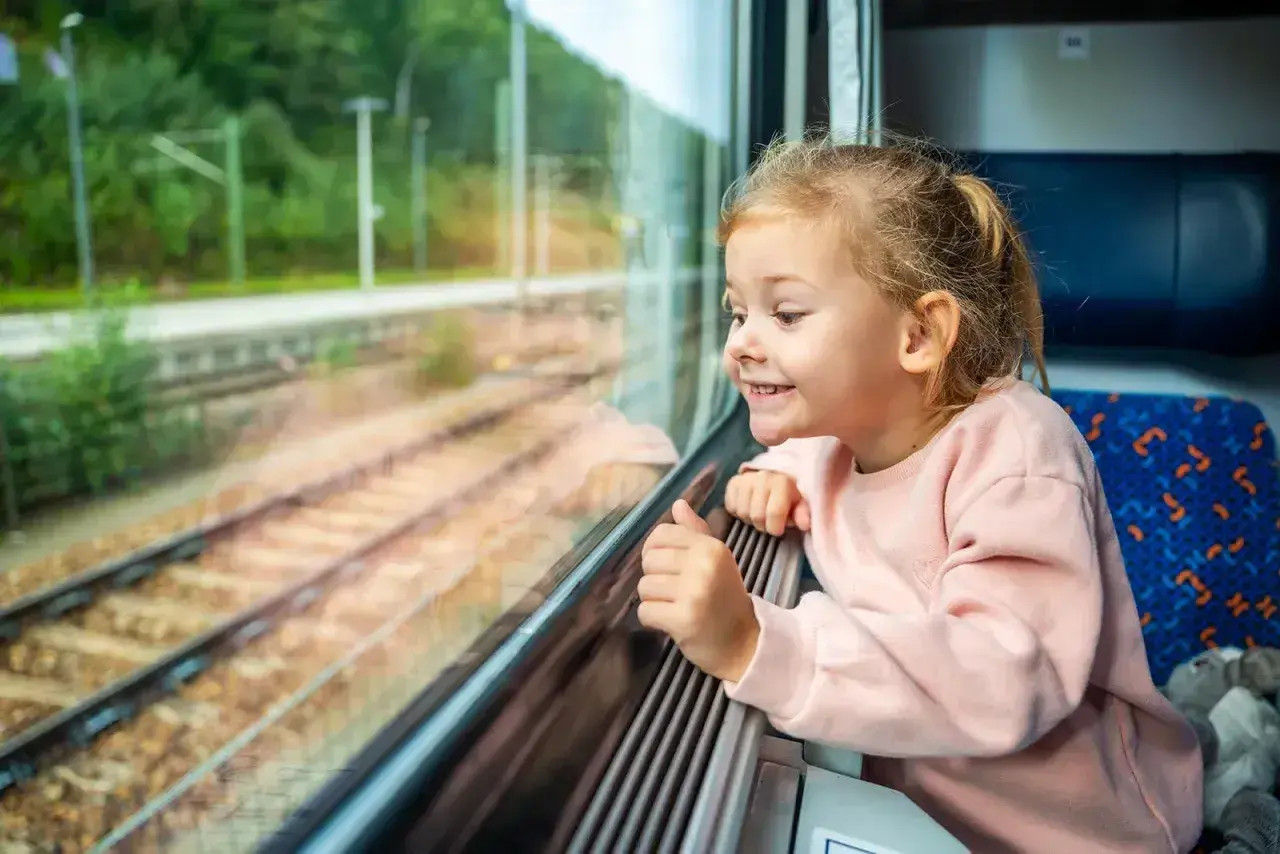 Uśmiechnięta dziewczynka patrzy przez okno pociągu. Zastanawia się, od ilu lat można jeździć samemu pociągiem, marząc o podróżach.