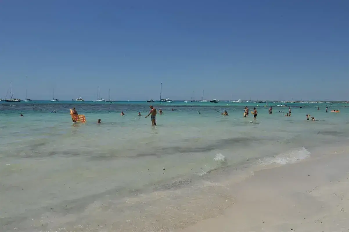Słoneczny dzień na plaży Es Trenc. Ludzie kąpią się w turkusowej wodzie, a na horyzoncie widać żaglówki.