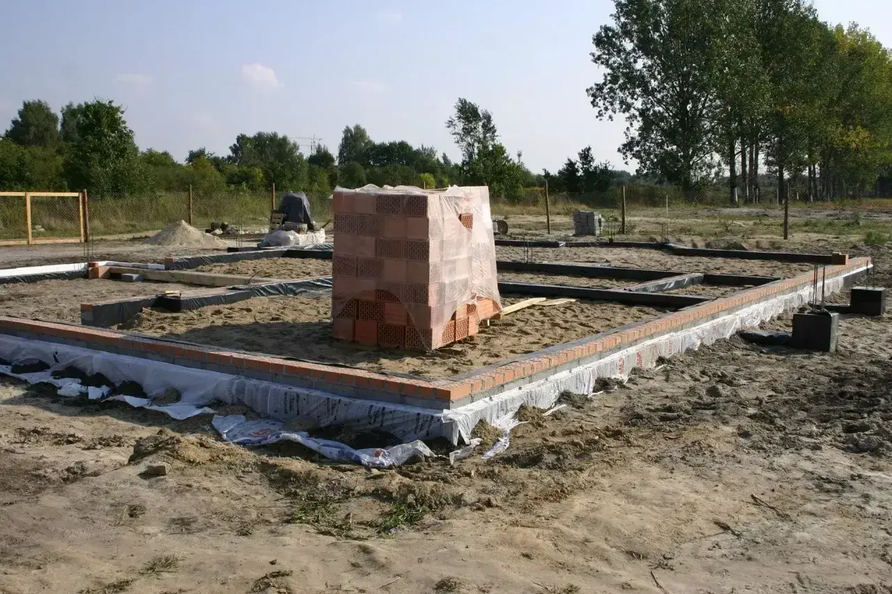 Stan zero budynku: fundamenty z cegły i betonu, piasek, folia izolacyjna, stos cegieł pod przykryciem.