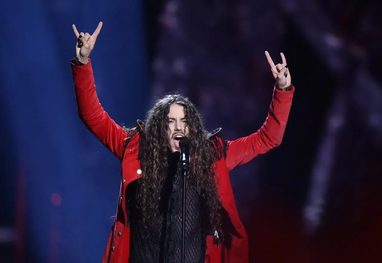 Michał Szpak na Eurowizji w czerwonej marynarce, z uniesionymi rękami i gestem rock'n'rolla.