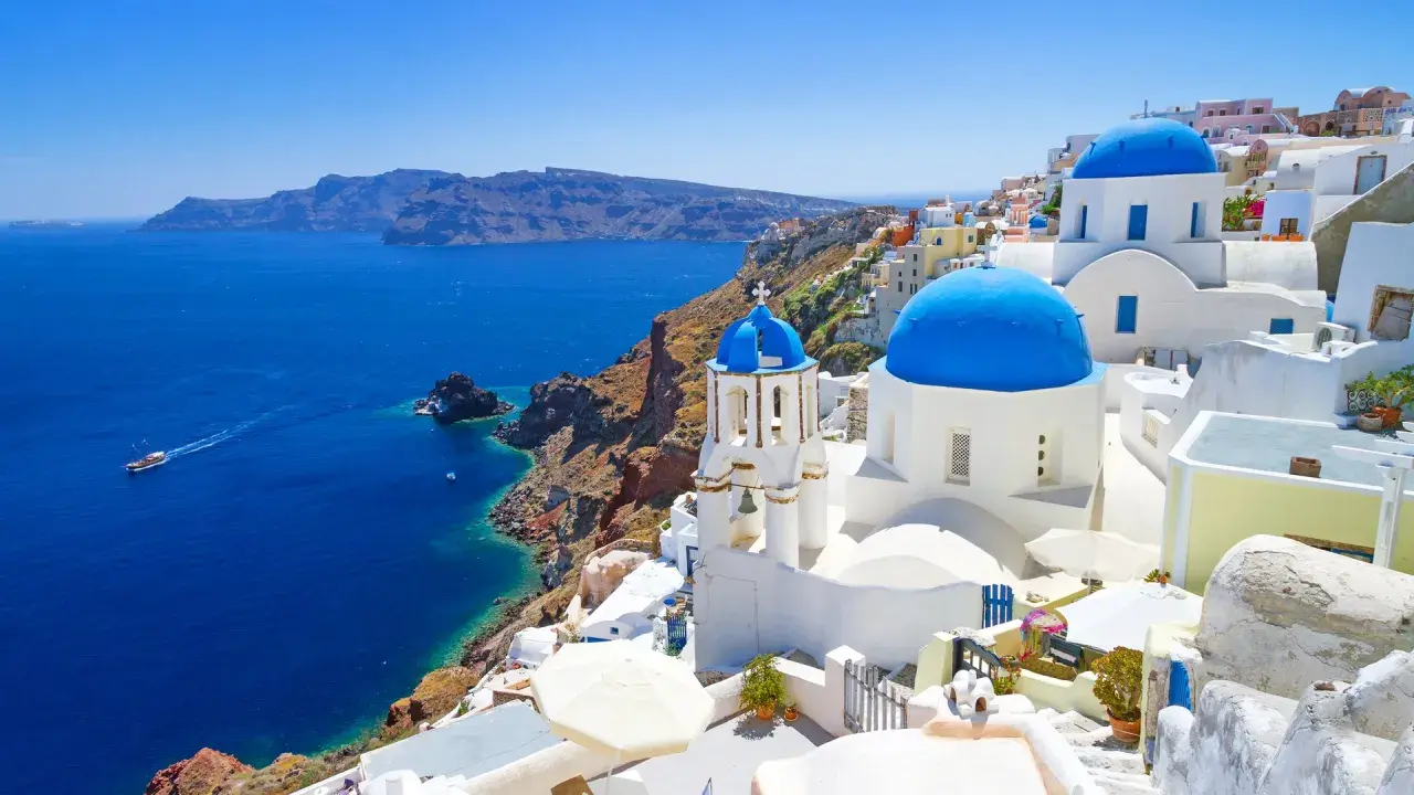 Santorini wycieczki: białe domy z niebieskimi kopułami na klifie nad błękitnym morzem. Statek płynie w oddali.