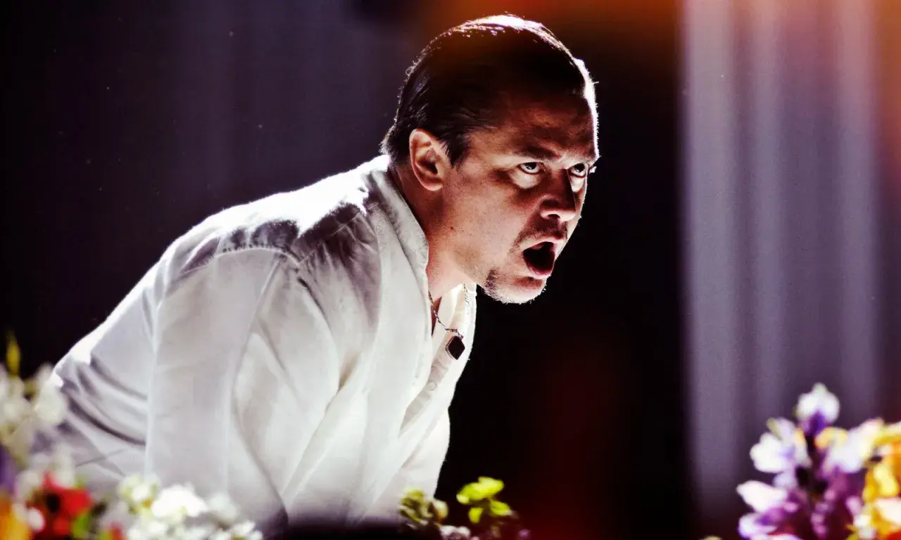Mike Patton heute, mit weit aufgerissenem Mund und intensivem Blick, trägt ein weißes Hemd. Blumen im Vordergrund.