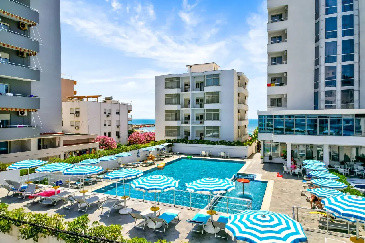 Albania - wczasy samolotem all inclusive. Basen hotelowy z leżakami i parasolami, w tle budynki i morze. Idealne miejsce na relaks.