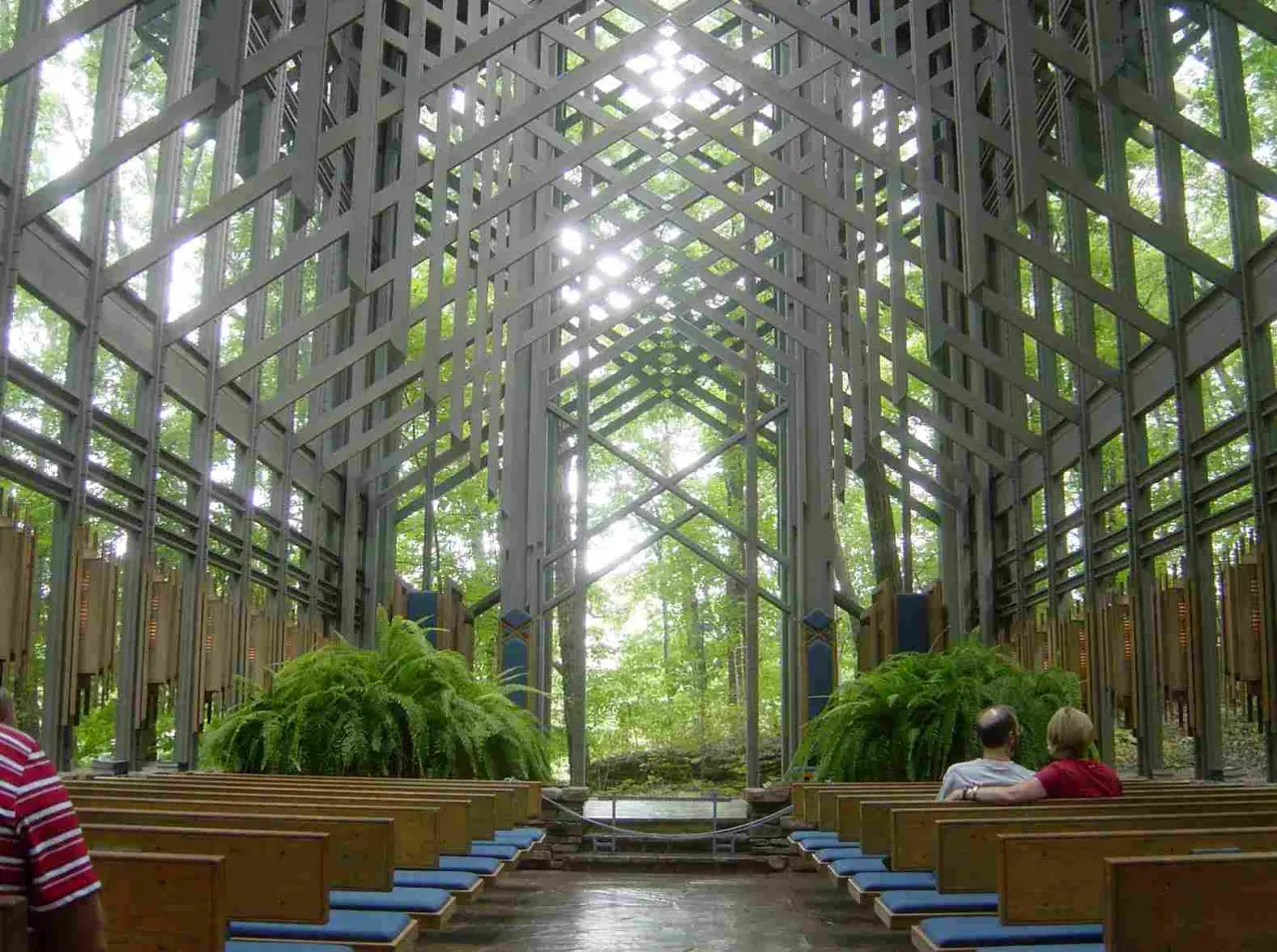 Wnętrze kaplicy w lesie, gdzie architektura organiczna łączy się z naturą. Drewniane ławki, paprocie i światło przenikające przez kratownicę tworzą spokojną atmosferę.