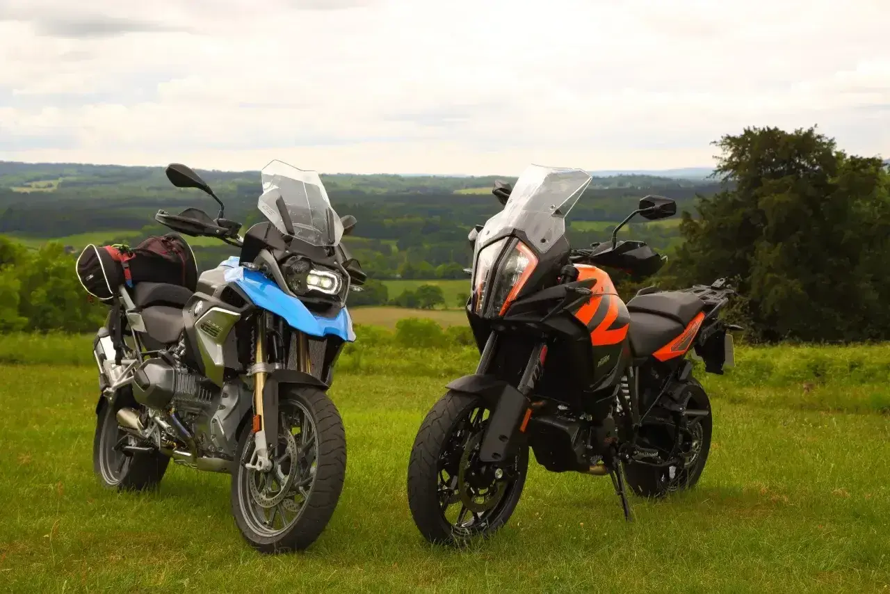 Dwa motocykle turystyczne, w tym pomarańczowy KTM 1290 Super Adventure, stoją na trawie z widokiem na zielone wzgórza.