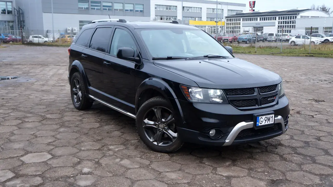 Czarny Dodge Journey 3.6 zaparkowany na placu z kostki brukowej. W tle budynki i inne samochody.