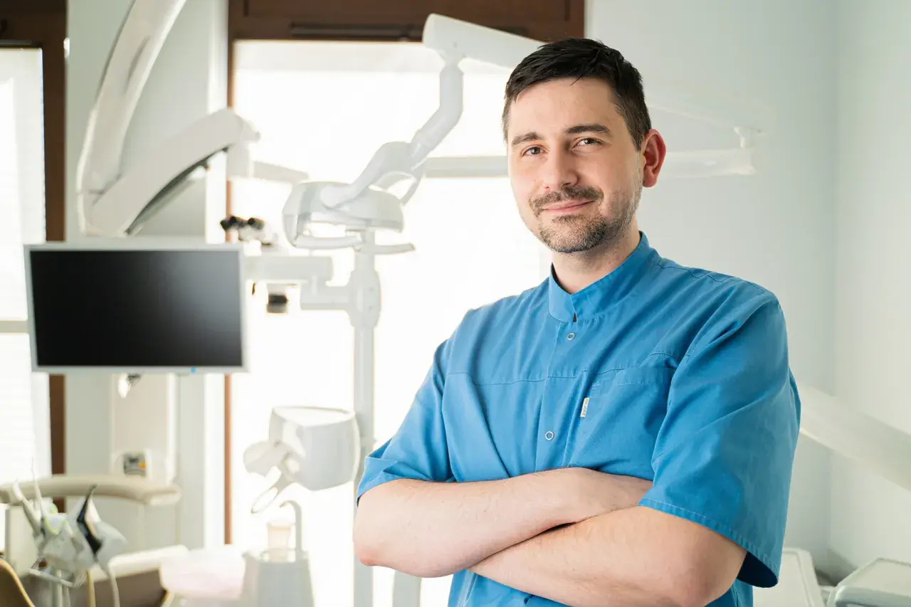 Uśmiechnięty dentysta w niebieskiej koszuli stoi ze skrzyżowanymi rękami w gabinecie stomatologicznym.