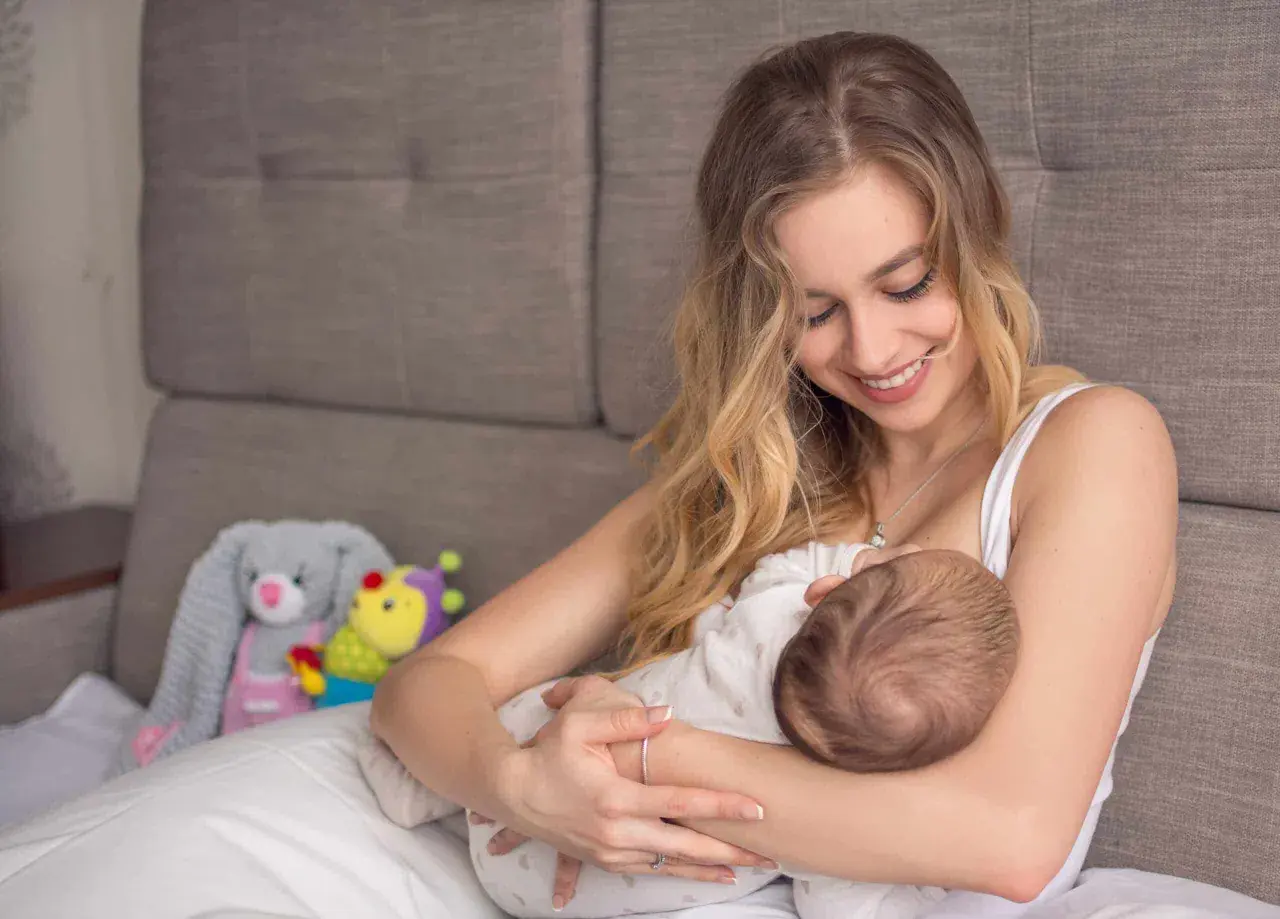 Uśmiechnięta mama karmi piersią dziecko. W tle pluszowe zabawki. Nakładki na pierś do karmienia mogą pomóc w tym intymnym momencie.