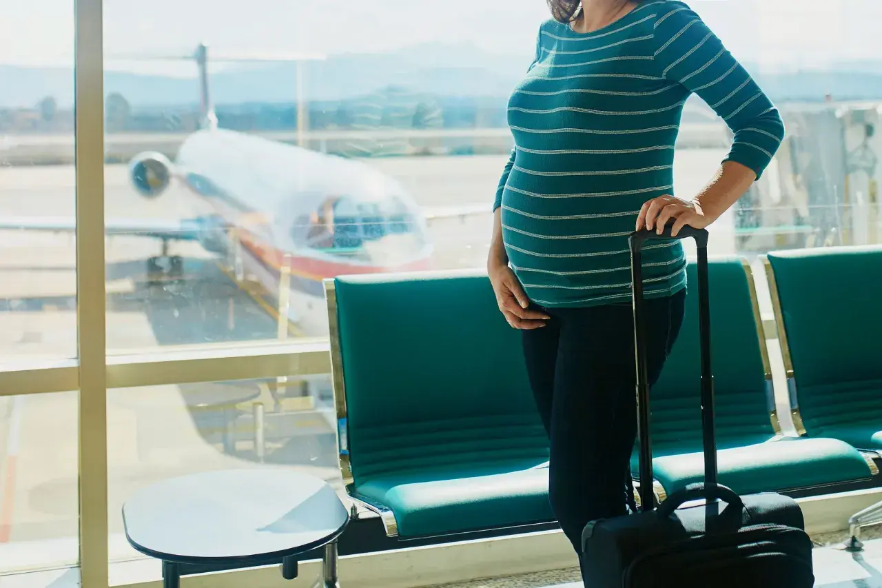 Ciężarna kobieta z walizką czeka na lotnisku. W tle widać samolot i pasażerskie fotele. Latanie samolotem w ciąży to wyzwanie, ale podróż dobiega końca.