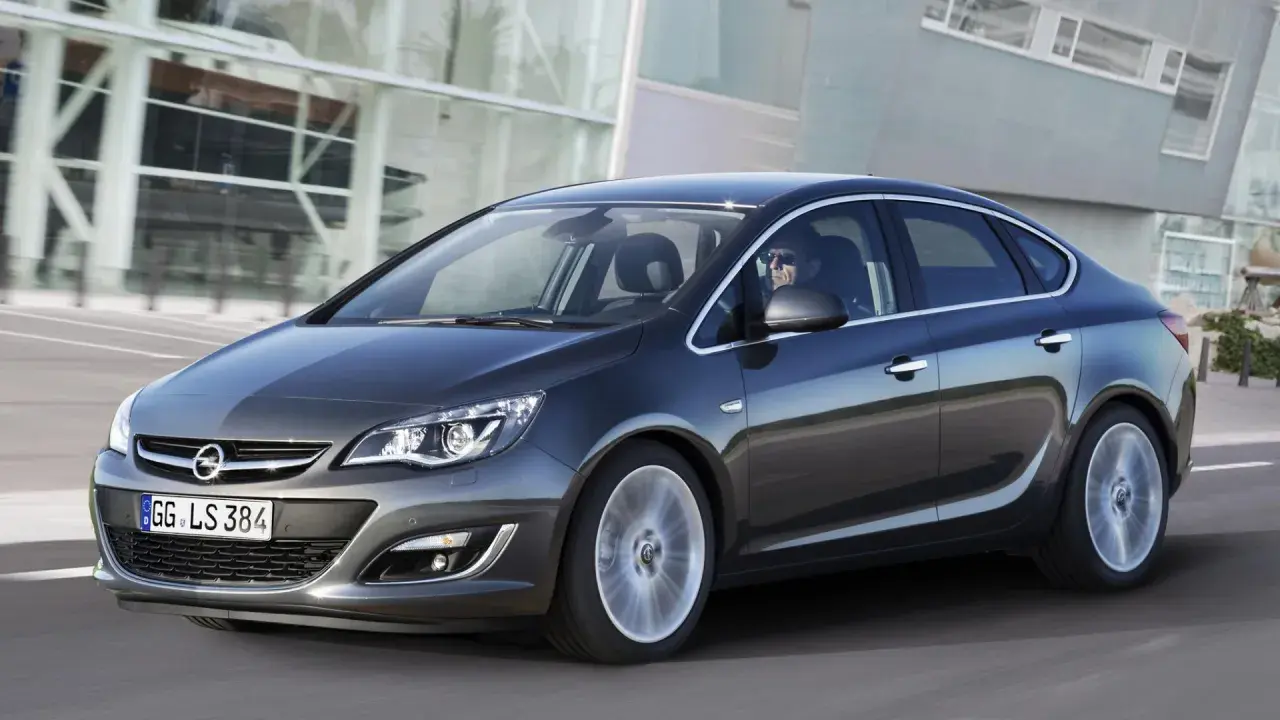 Ciemnoszary Opel Astra J 1.6 benzyna w ruchu, z kierowcą w okularach za kierownicą, na tle nowoczesnego budynku.
